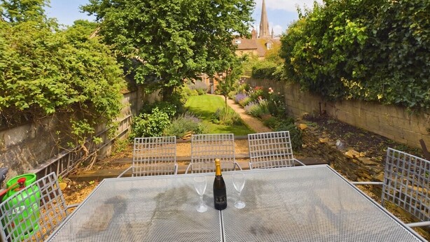 A patio with a table, chairs, and a bottle of champagne. There is a garden with a lawn and a building with a spire in the background. - Cosy Cottage in Moreton-in-Marsh (Gloucestershire)