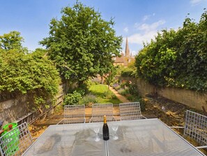 A patio with a table, chairs, and a bottle of champagne. There is a garden with a lawn and a building with a spire in the background.