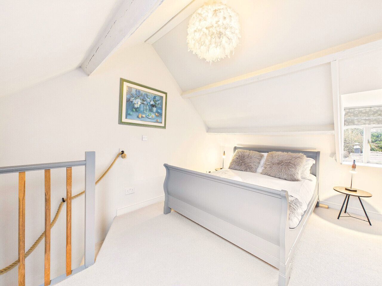 A bedroom with a bed, a small table, a painting, and a window.