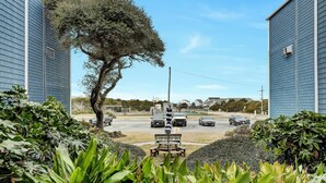 A view of a parking area with a bench and a tree in the foreground, surrounded by greenery. - Captain's Quarters | Oceanfront Condo (North Topsail Beach)