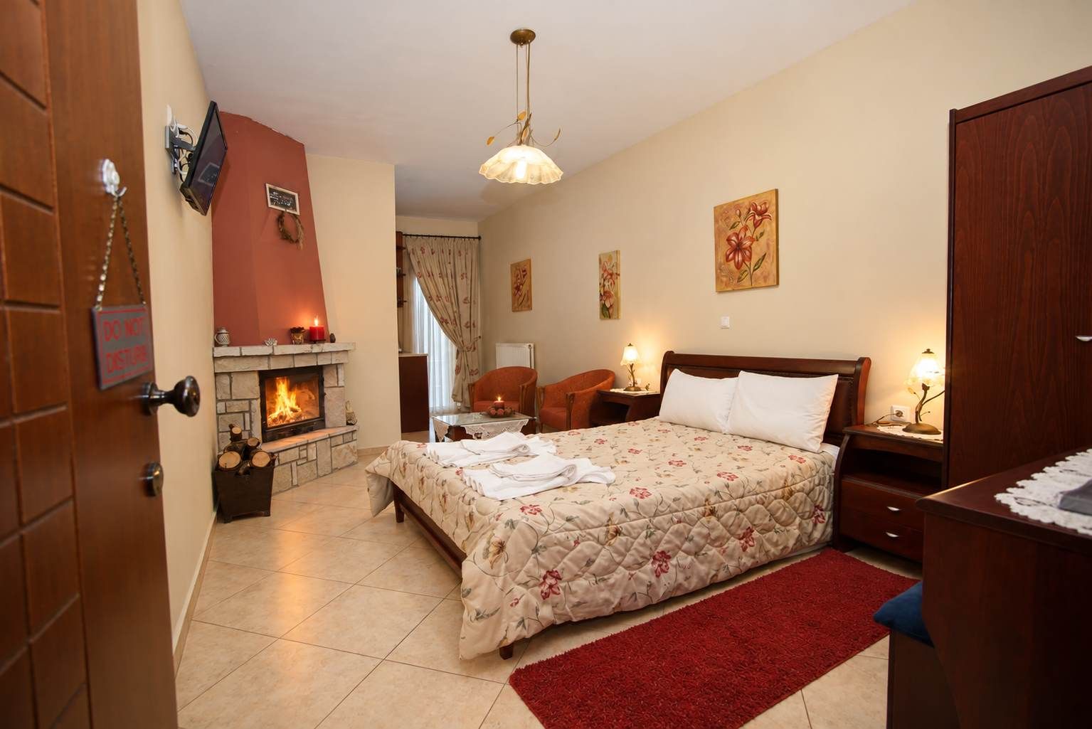 A hotel room with a fireplace, a bed with a floral bedspread, a red rug, and a nightstand with a lamp.