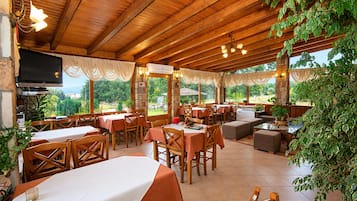 An outdoor restaurant with wooden tables and chairs, a flat-screen TV, and a view of the outdoors.