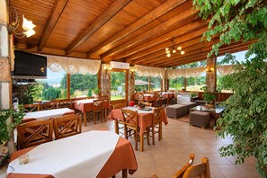 An outdoor restaurant with wooden tables and chairs, a flat-screen TV, and a view of the outdoors.