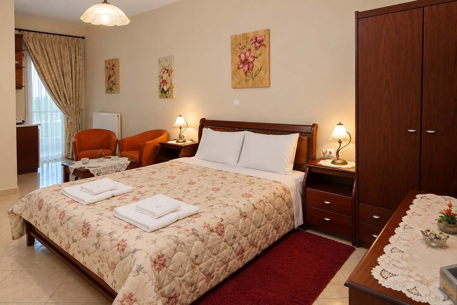 A hotel room with a large bed, two bedside tables with lamps, a wooden wardrobe, a red rug, and floral artwork on the walls.