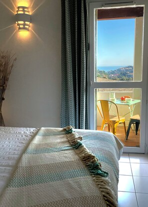 A bedroom with a bed, a view of the outdoors through a sliding door, and a table with chairs outside. - Les Vigies -T4 vue mer-terrasse-parking-6 pers (Collioure)