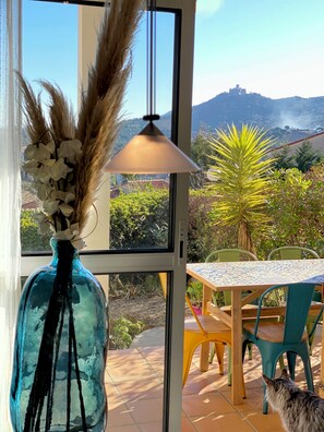 Quarto com uma janela grande, um vaso com flores brancas, uma mesa com cadeiras e vista para uma colina com edifícios.