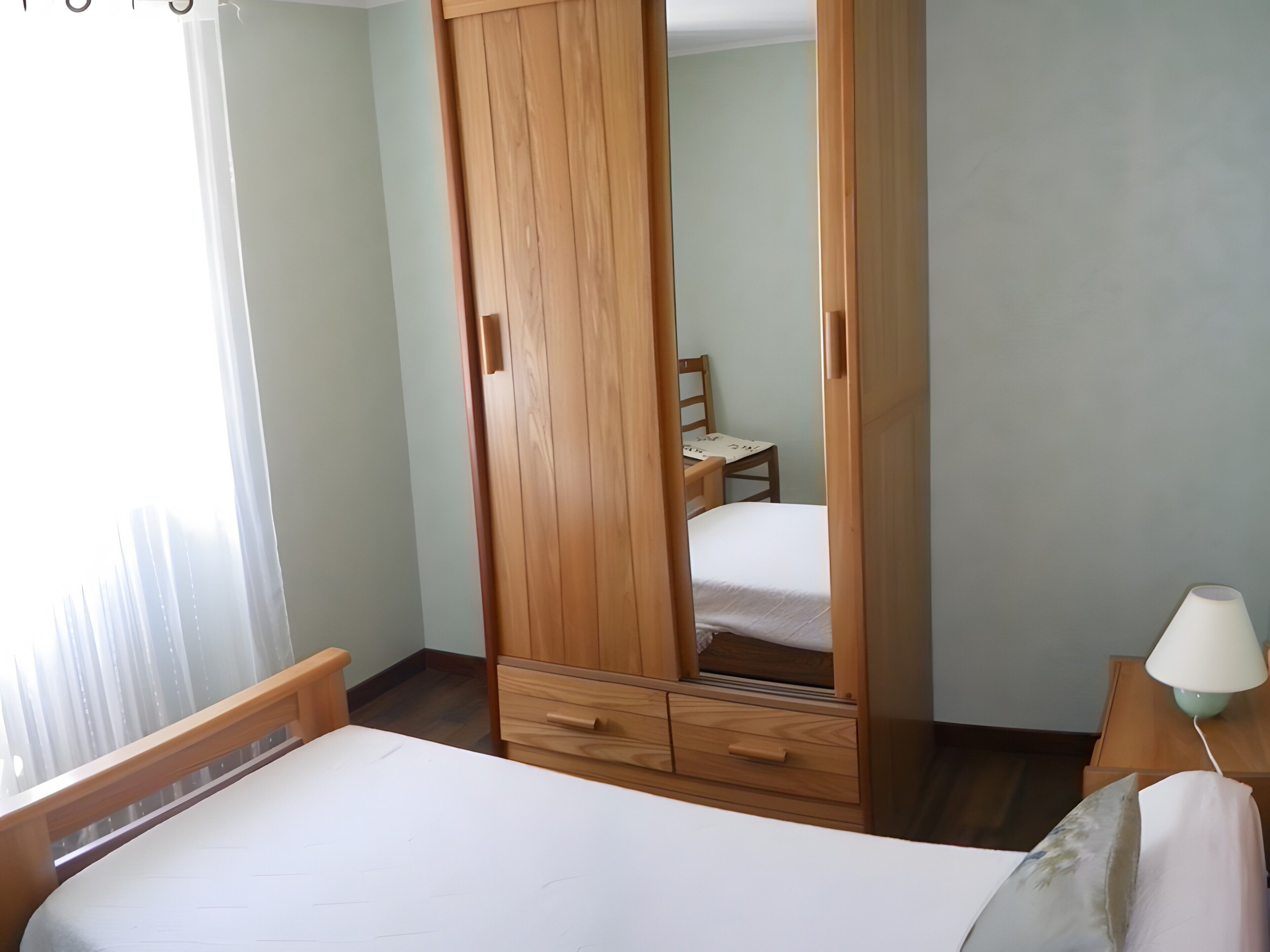 A bedroom with a bed, wooden wardrobe, a chair, a nightstand with a lamp, and a window with curtains.