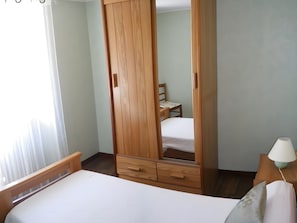 Een slaapkamer met een bed, een houten garderobe, een stoel, een nachtkastje met een lamp en een raam met gordijnen.