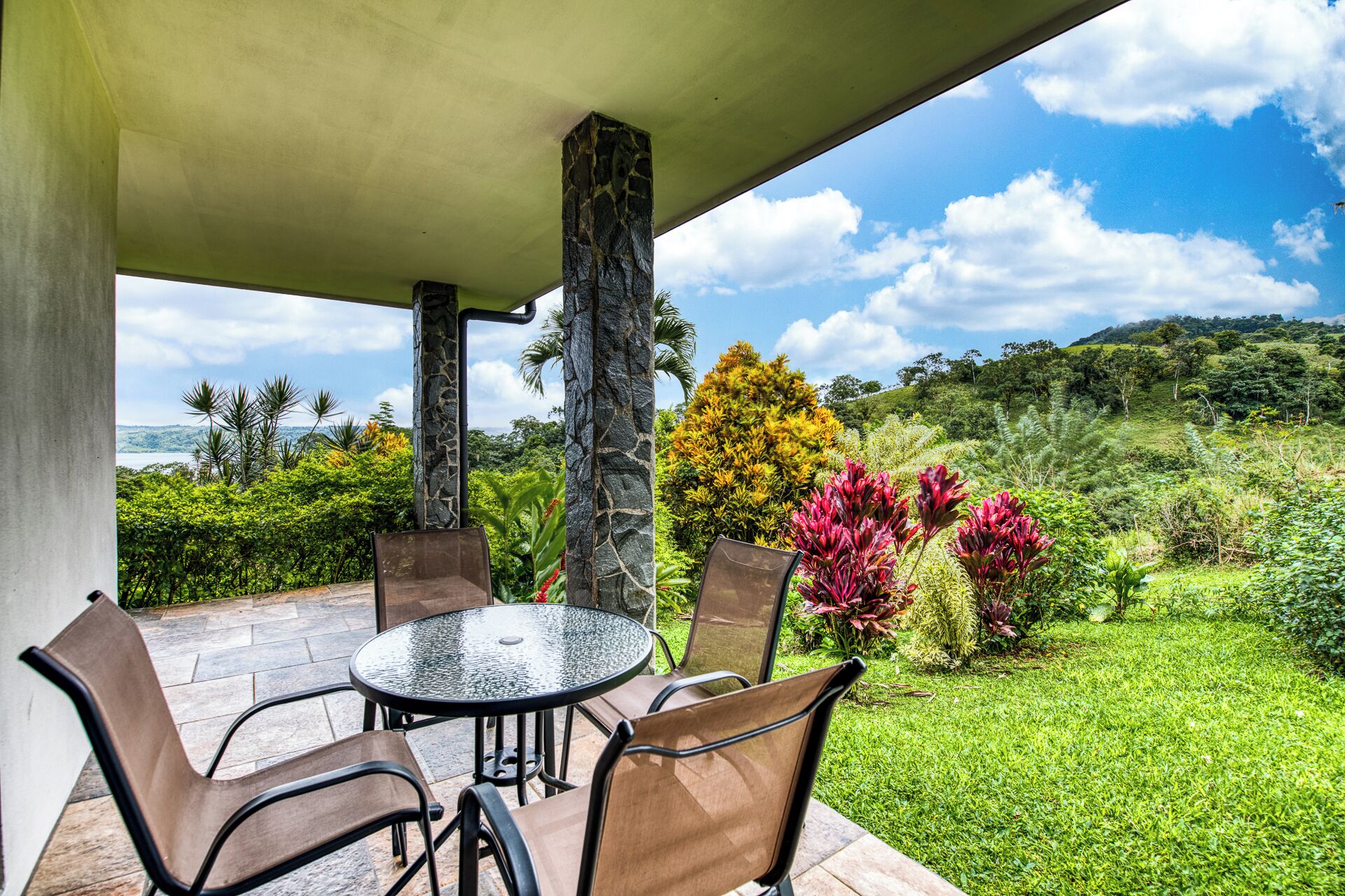An outdoor patio with a round table and chairs, surrounded by lush greenery and a view of the ocean.