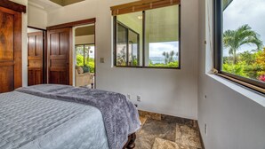 A bedroom with a large bed, a wooden door, a window with a view of greenery, and a mirror on the wall.