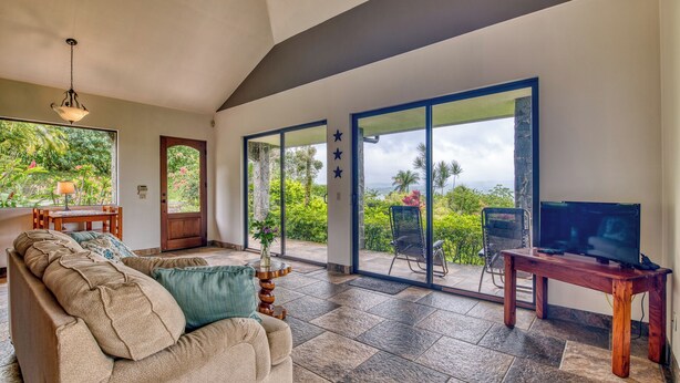 A spacious living area with a large sofa, a television, and a sliding glass door leading to a balcony with a view of lush greenery and the ocean.