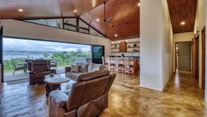 Amplia sala de estar con un gran ventanal que da a un paisaje pintoresco, un rincón de descanso con sofás de cuero y una cocina moderna con armarios de madera.