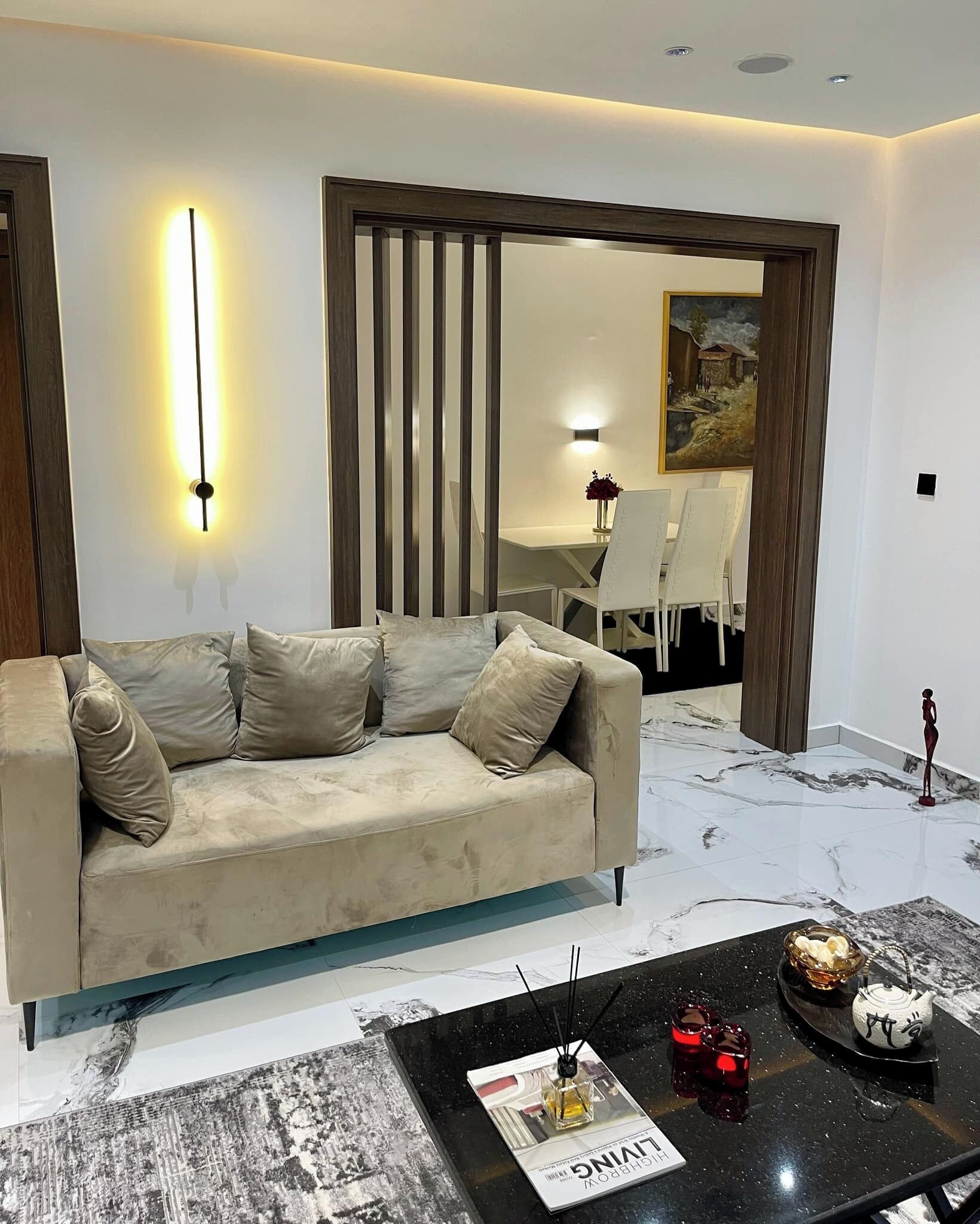 A modern living room with a beige sofa, a black coffee table with magazines and decorative items, and a marble floor.