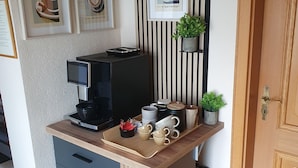 Une petite cuisine équipée avec une machine à café, des tasses et un plateau. Les murs sont ornés de tableaux encadrés et une petite étagère est décorée de plantes.