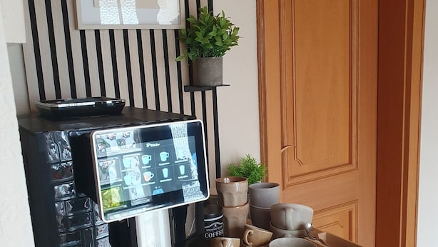 Une machine à café dotée d’un écran numérique, un panneau décoratif représentant un expresso, une petite plante en pot et un plateau avec des tasses à café et une boîte.