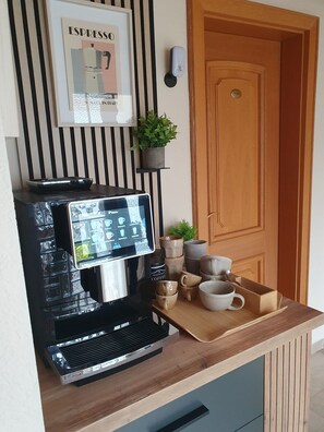 Une machine à café dotée d’un écran numérique, un panneau décoratif représentant un expresso, une petite plante en pot et un plateau avec des tasses à café et une boîte.
