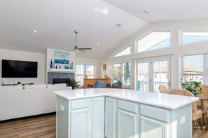 A spacious kitchen with a large island, a dining area with wicker chairs, and a flat-screen TV mounted on the wall. - True Island Retreat in the Heart of Kure Beach! (Kure Beach)