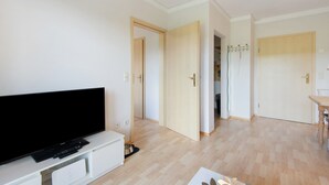 Una sala de estar moderna con un televisor de pantalla plana, un mueble blanco para entretenimiento, piso de madera y un comedor con mesa y sillas.