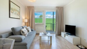Una sala de estar con sofá, mesa de centro, televisor de pantalla plana y vista a un campo verde a través de amplias ventanas.