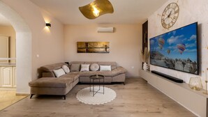 A living room with a sectional sofa, a round coffee table, a white rug, a wall-mounted clock, and a large TV screen displaying a landscape scene. - Solitude in Ružići - Istrien / Solitude in Ružići - Appartement für 4 Personen (Ružići)