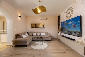 Ein Wohnzimmer mit einem Ecksofa, einem runden Couchtisch, einem weißen Teppich, einer Wanduhr und einem großen Fernsehbildschirm, der eine Landschaft zeigt.