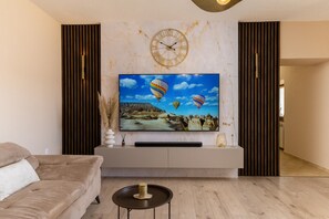 Ein modernes Wohnzimmer mit einem großen Flachbildfernseher an einer weißen Konsole, einem beigefarbenen Sofa und Holzböden.