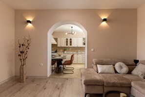 Ein modernes Wohnzimmer mit einem beige-farbenen Sofa, einem Essbereich mit Tisch und Stühlen sowie einer durch einen Bogen sichtbaren Küche.