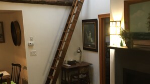 Una sala de estar con una escalera de madera apoyada en la pared, una chimenea, un comedor con sillas y una escalera que lleva a un nivel superior.