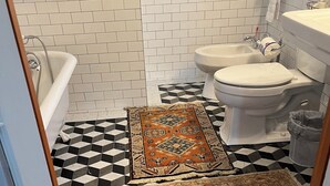 Un baño con bañera, inodoro y lavamanos, con piso a cuadros blanco y negro y alfombras estampadas.