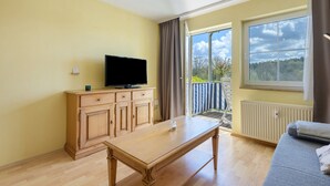 A room with a wooden table, a television on a cabinet, a sofa, and a large window with a view of trees. - Superior Apartment with Dog – Lake View, Balcony, Self Check-in (Spalt)