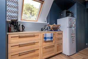 Een keuken met een raam, houten kastjes, een witte koelkast en een magnetron erboven.