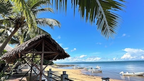 A wooden deck with a thatched roof structure, overlooking a beach with boats and a clear blue sky. - Holiday Beach Hotel (Nosy Be)