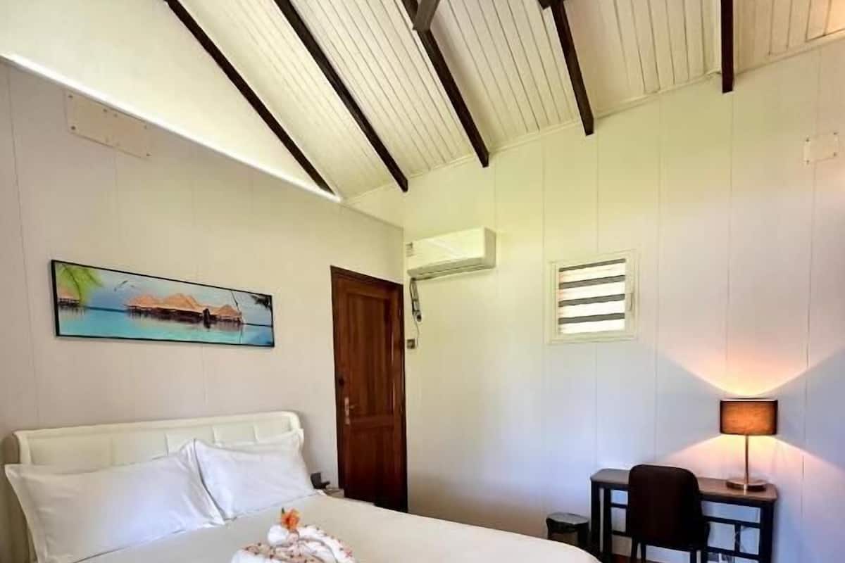 A hotel room with a bed, a desk, a chair, a painting, and a ceiling fan.