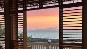A dining area with a table and chairs, large windows with wooden blinds, and a view of the sea and mountains. - Holiday Beach Hotel (Nosy Be)