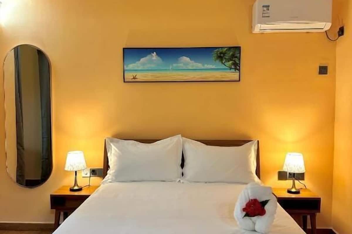 A hotel room with a bed, two bedside lamps, a wall-mounted air conditioner, a painting of a beach scene, and a mirror on the wall.