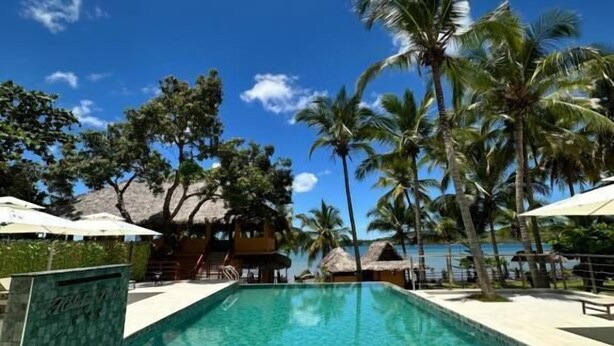 A clear blue pool surrounded by palm trees and thatched-roof structures, with a clear sky above. - Holiday Beach Hotel (Nosy Be)