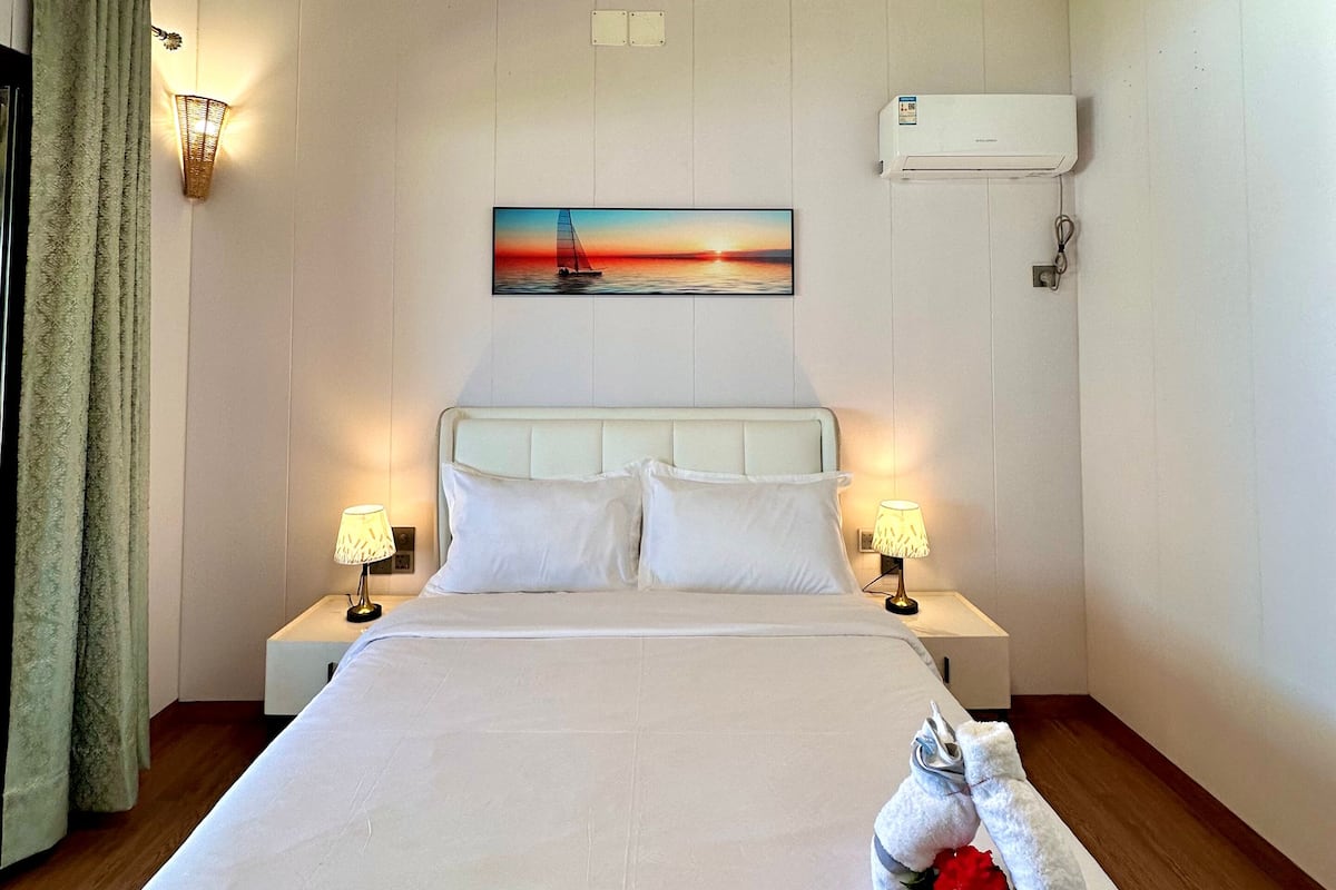 A hotel room with a large bed, two bedside tables with lamps, a ceiling fan, an air conditioning unit, and a painting of a sunset over water on the wall.