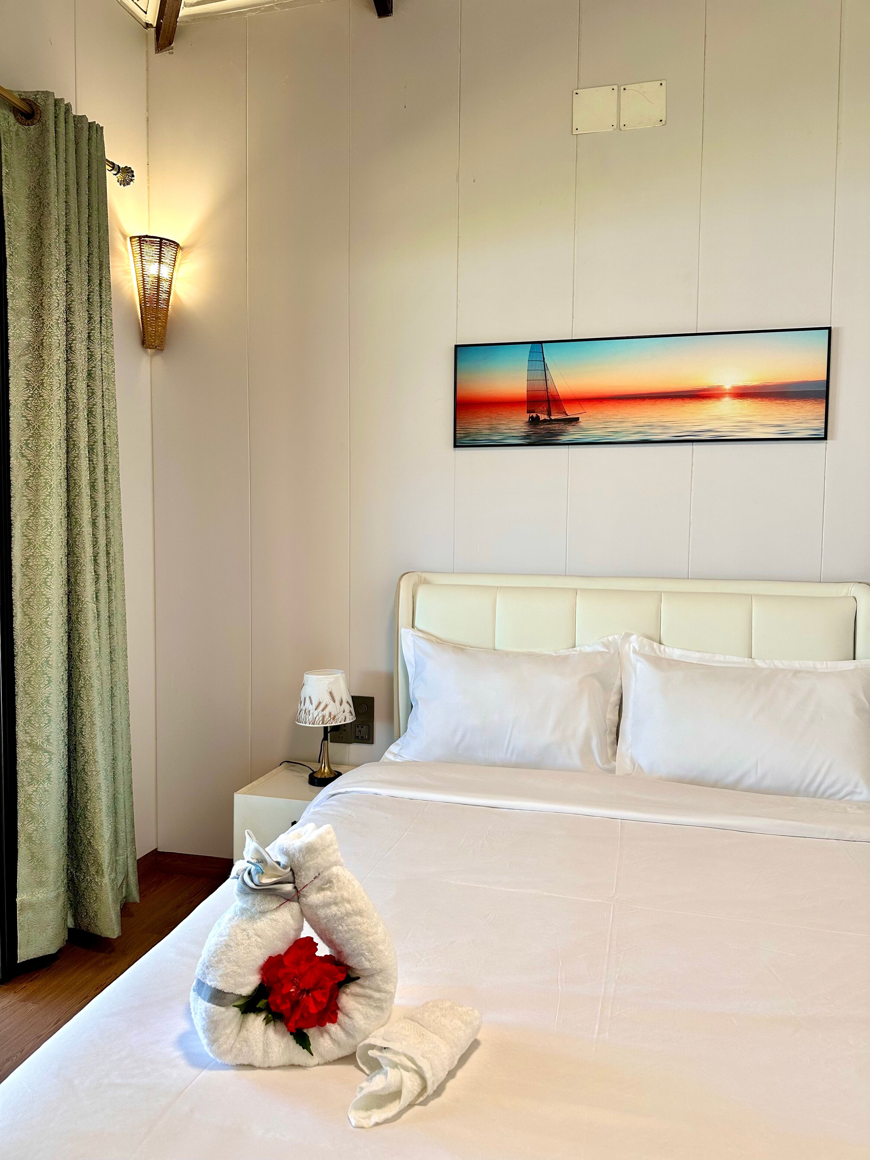 Una camera d'albergo con un letto, un quadro raffigurante un tramonto sull'acqua, un comodino con una lampada e un asciugamano con un fiore sul letto.