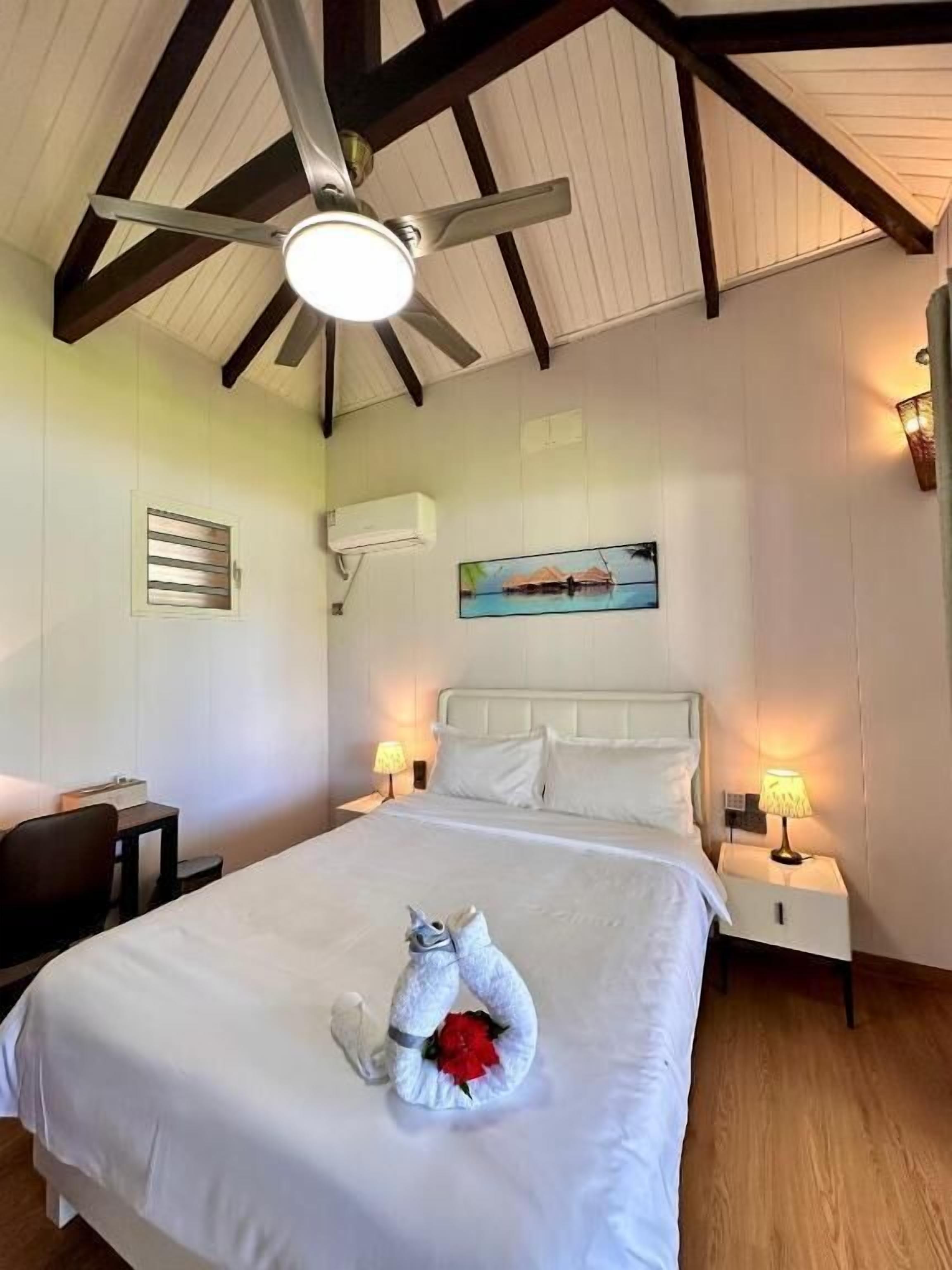 Quarto de hotel com cama, escrivaninha, cadeira, ventoinha no teto e duas luminárias de cabeceira.