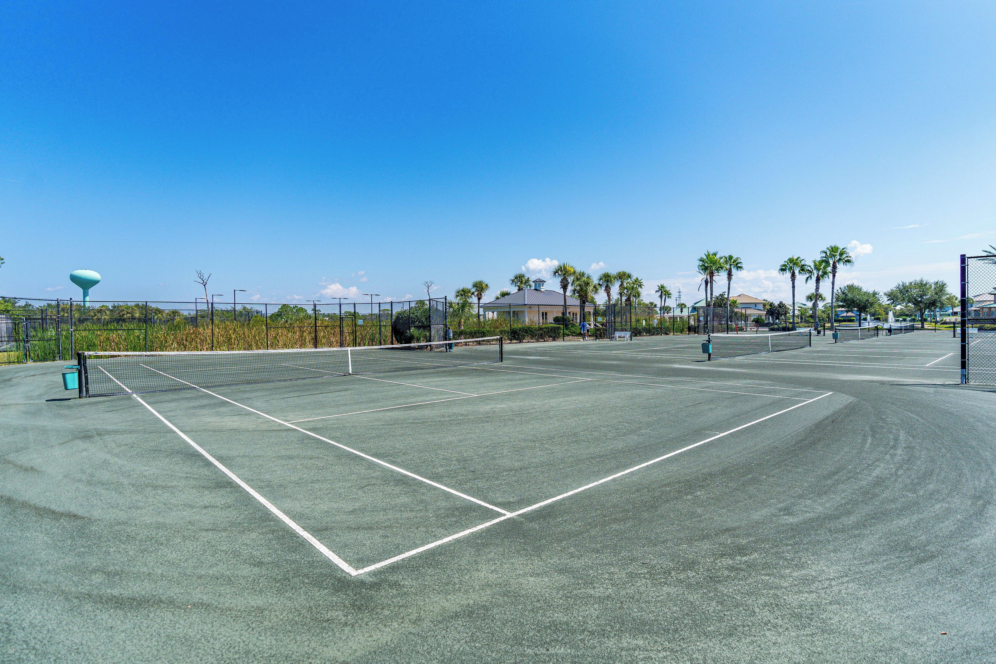 A tennis court with a net, surrounded by a fence, palm trees, and a building in the background.