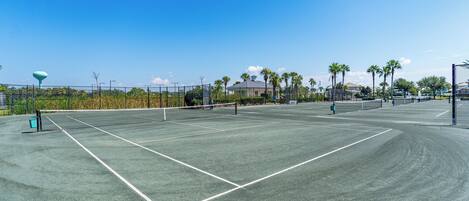 A tennis court with a net, surrounded by a fence, palm trees, and a building in the background.