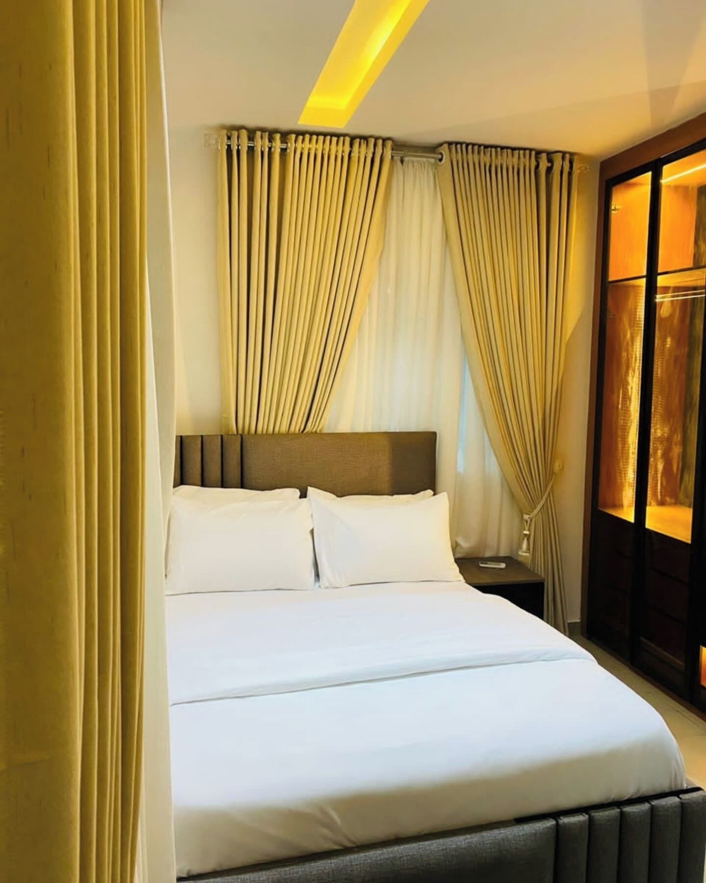 Ein Hotelzimmer mit Bett, Vorhängen, einem Nachttisch und einem Fenster mit Vorhängen.