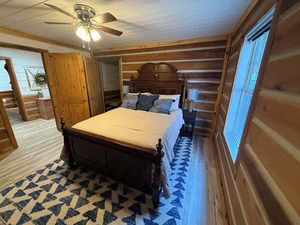 Un dormitorio de madera con una cama, un mesita de noche, una lámpara, un ventilador de techo y una ventana.