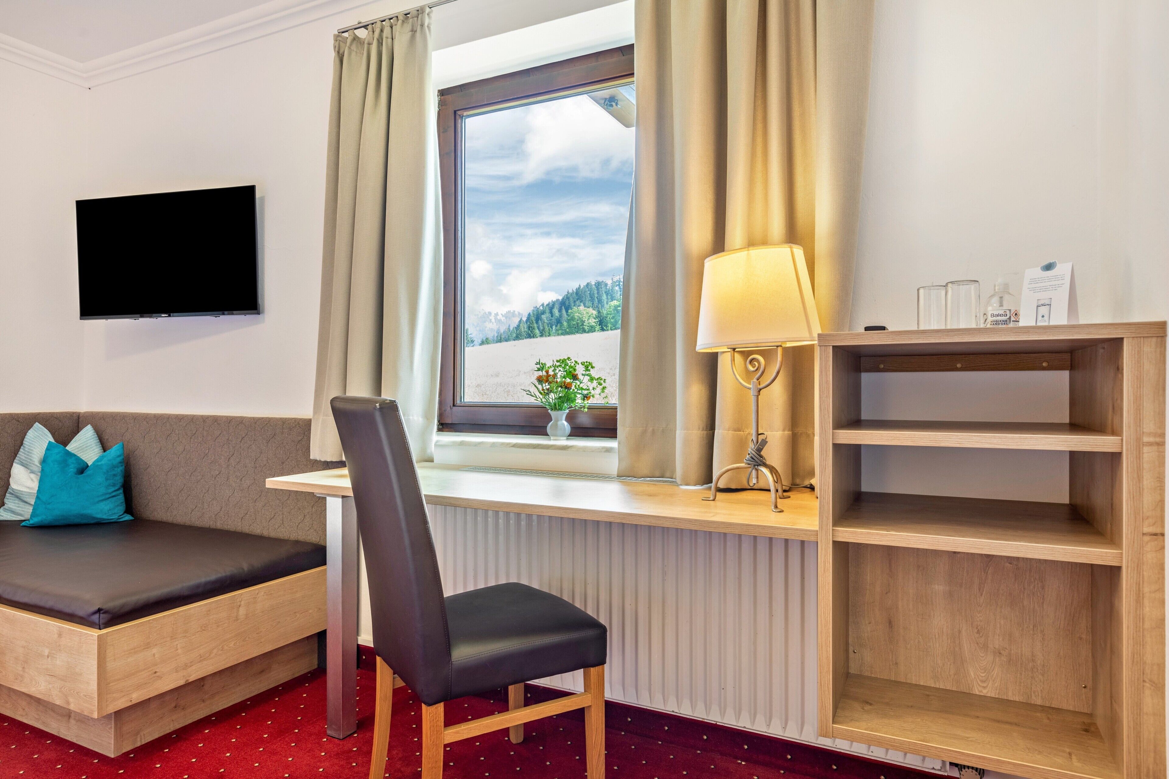 A room with a bed, a desk with a lamp, a TV, and a window with curtains.