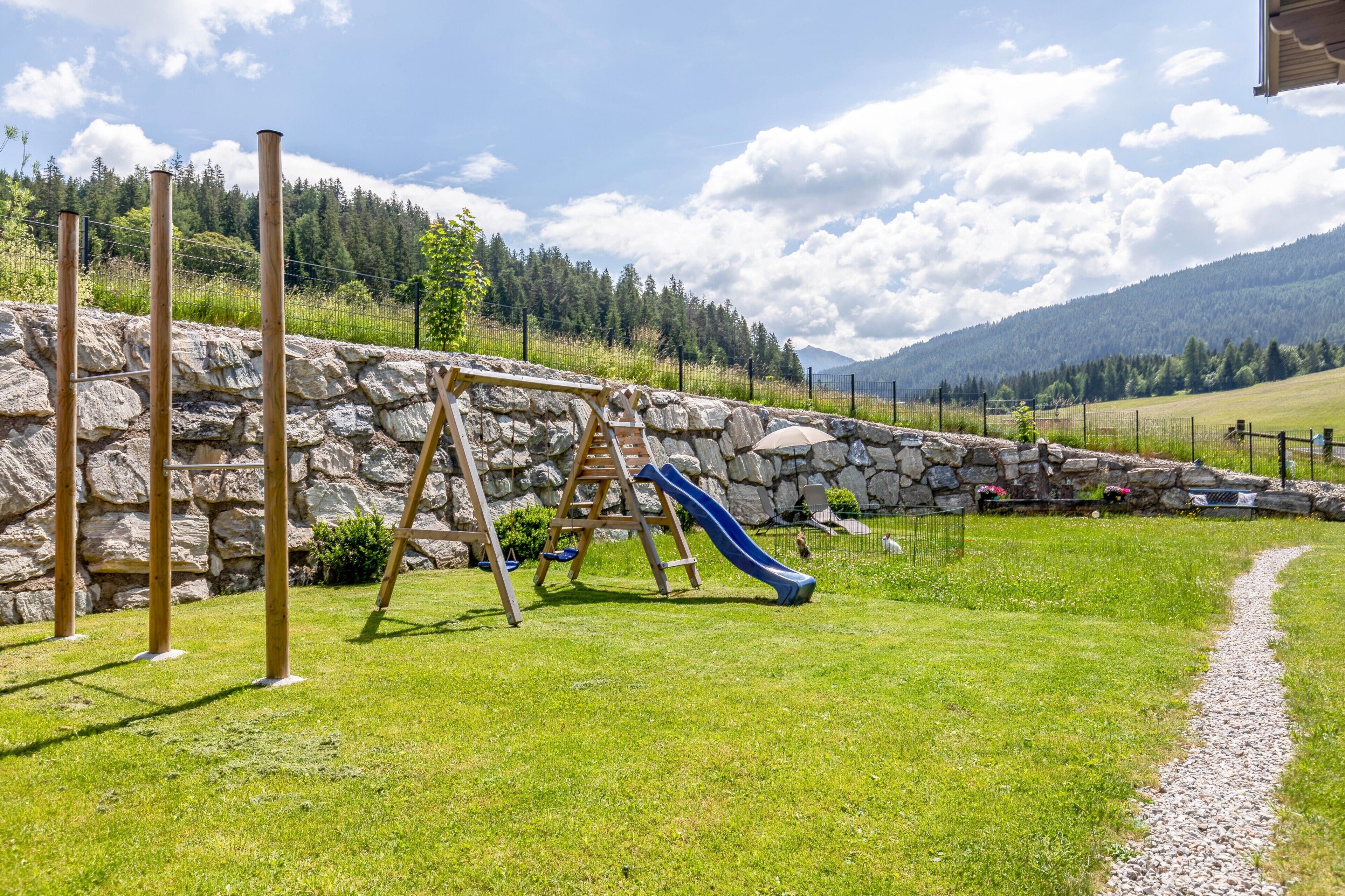 A playground with a slide and swings, surrounded by a stone wall and a grassy area.