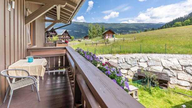 A wooden balcony with a table and chair, a stone wall, and a view of a mountain landscape.