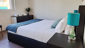 Un lit bien fait, recouvert d’une couverture bleue, une table de chevet avec une lampe turquoise et une petite plante en pot posée sur une commode.