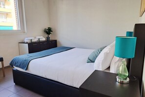 A neatly made bed with a blue blanket, a bedside table with a teal lamp, and a small potted plant on a dresser. - Close to the Heart of Lugano (Mola Glicine) (Lugano)