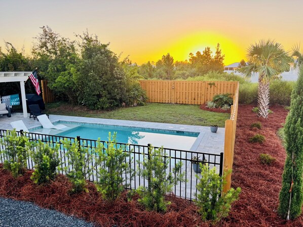 Ein Garten mit Schwimmbad, einer Holzterrasse und einem eingezäunten Bereich mit verschiedenen Pflanzen.