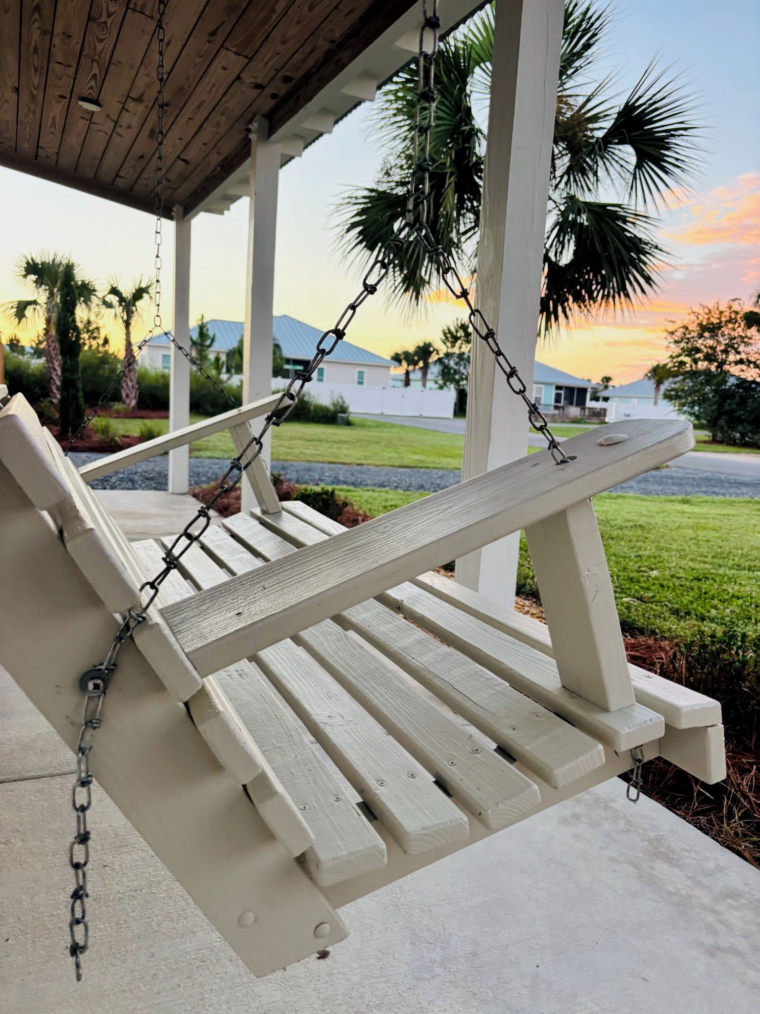 Una poltrona a dondolo in legno sospesa da un portico tramite una catena, con vista su una zona residenziale con case e palme.